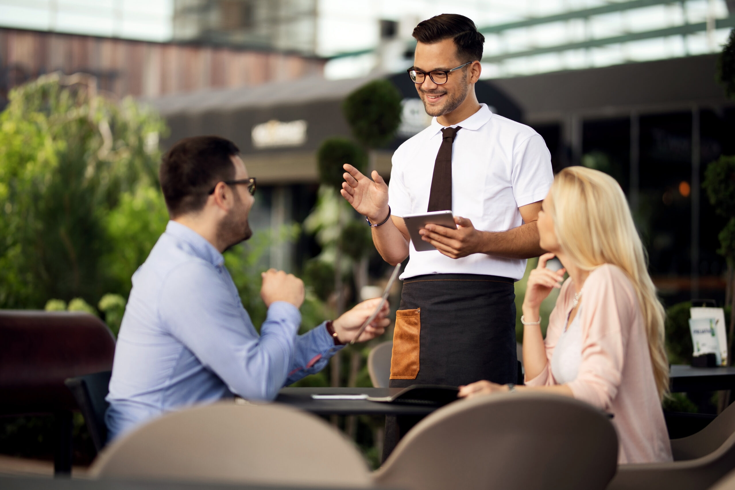 Young happy waiter using digital tablet while talking to a couple and taking their order in a cafe,